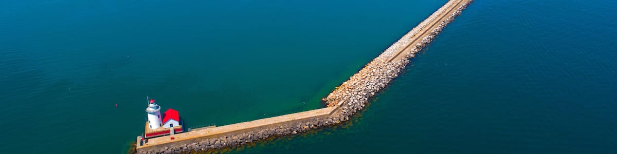 Ein Leuchtturm auf einem langen Steg, der sich in ein großes Gewässer erstreckt, umgeben von einer kleinen Insel mit Gebäuden und Grünflächen.