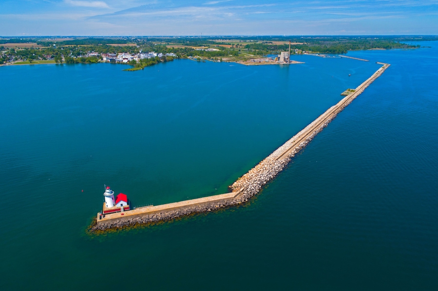 A lighthouse on a long pier extending into a large body of water, surrounded by a small island with buildings and greenery.