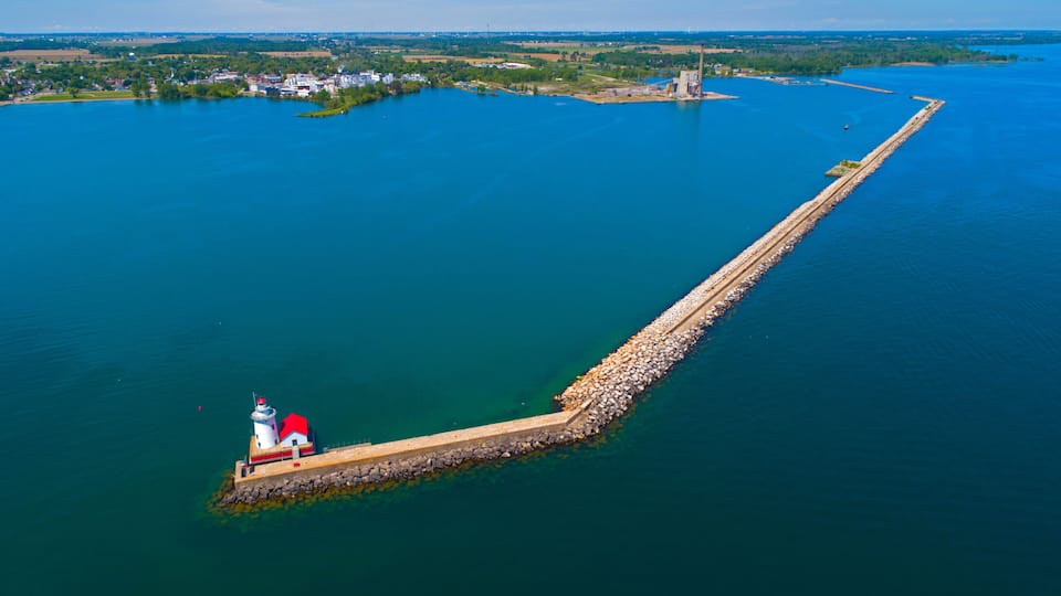 A lighthouse on a long pier extending into a large body of water, surrounded by a small island with buildings and greenery.