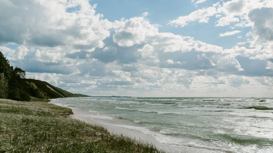 A beach with waves, grassy dunes, and a cloudy sky.