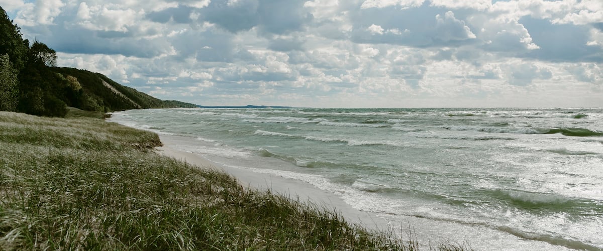 A beach with waves, grassy dunes, and a cloudy sky.