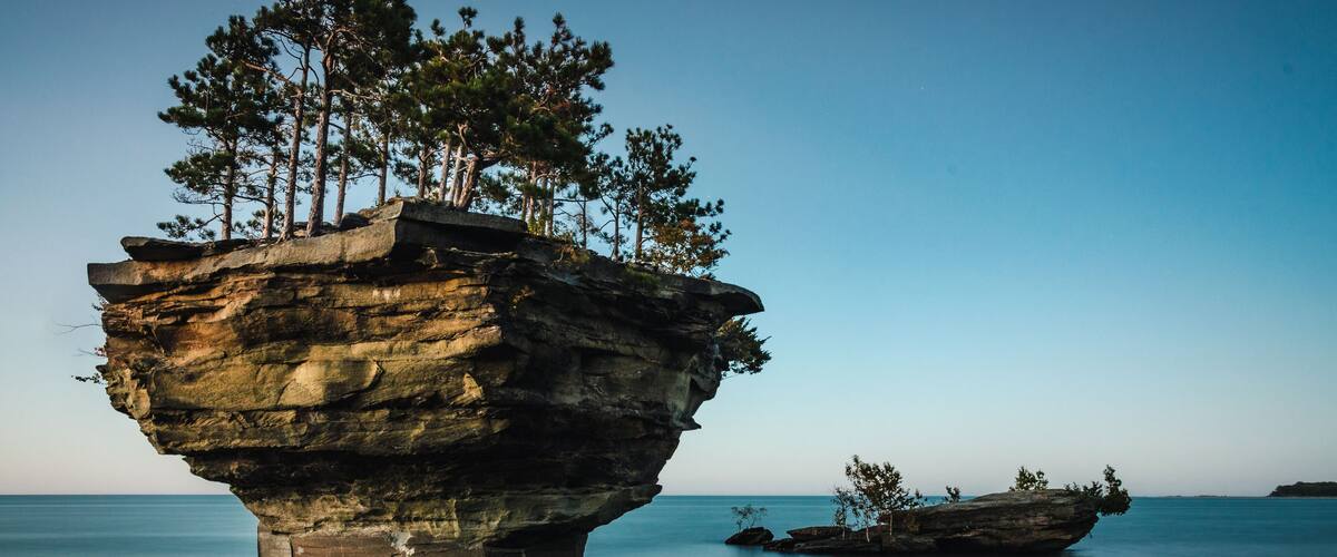 A rock formation with trees on top, surrounded by calm water.
