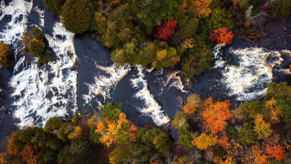 Incredible panoramic travel aerial look down photograph of Bond Falls Scenic Site including the upper and lower waterfalls and rapids in autumn with fall colored foliage on the treetops below.