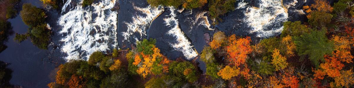 Incredible panoramic travel aerial look down photograph of Bond Falls Scenic Site including the upper and lower waterfalls and rapids in autumn with fall colored foliage on the treetops below.