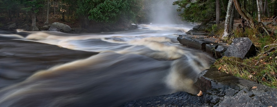 A fast-flowing river with a waterfall, surrounded by rocks and trees.