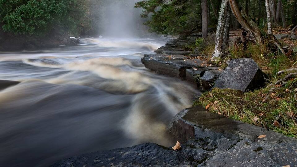 A fast-flowing river with a waterfall, surrounded by rocks and trees.