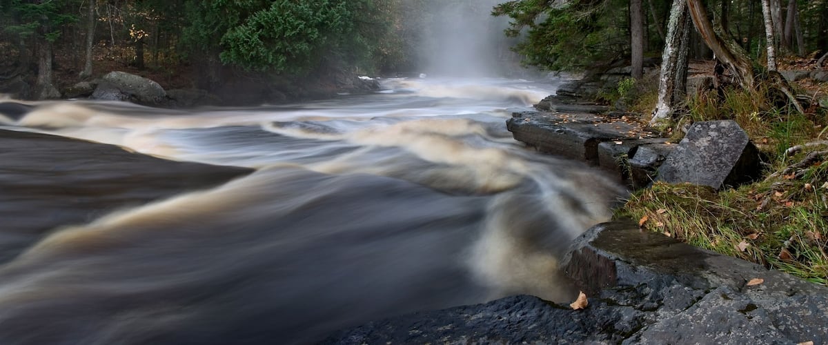 A fast-flowing river with a waterfall, surrounded by rocks and trees.