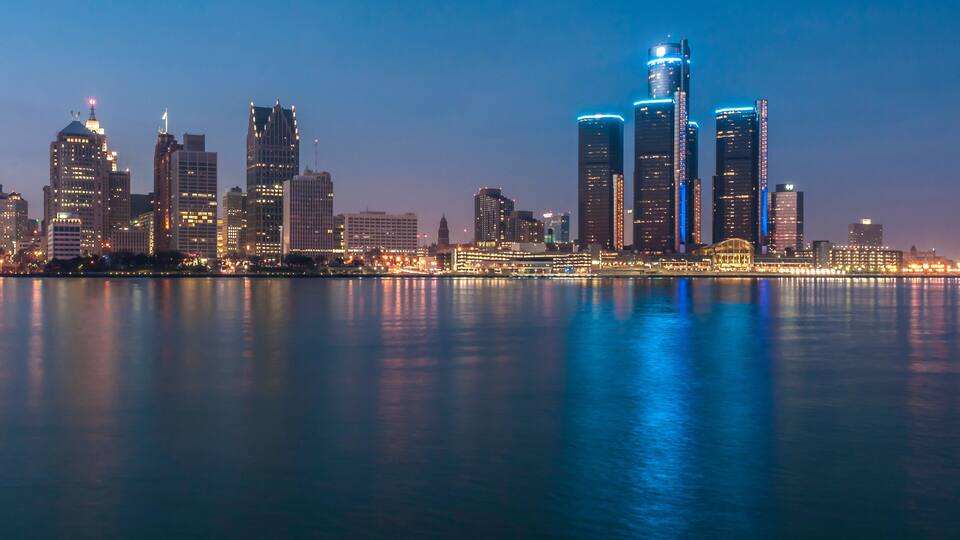 A city skyline at dusk with illuminated buildings reflecting on the water.