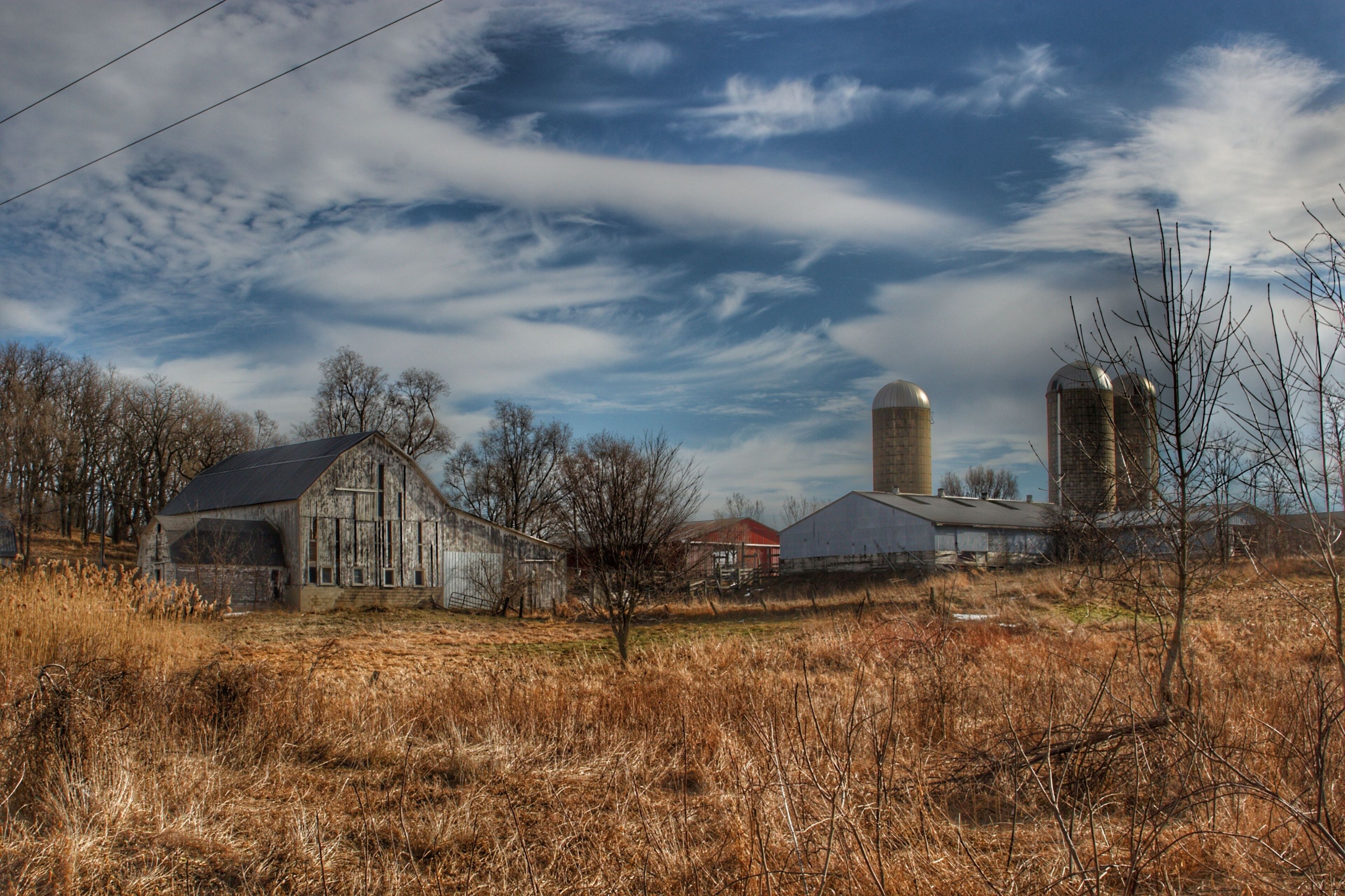 A rural landscape with a barn, silo, and farm buildings under a partly cloudy sky.