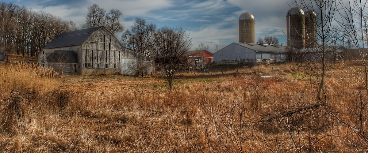 A rural landscape with a barn, silo, and farm buildings under a partly cloudy sky.