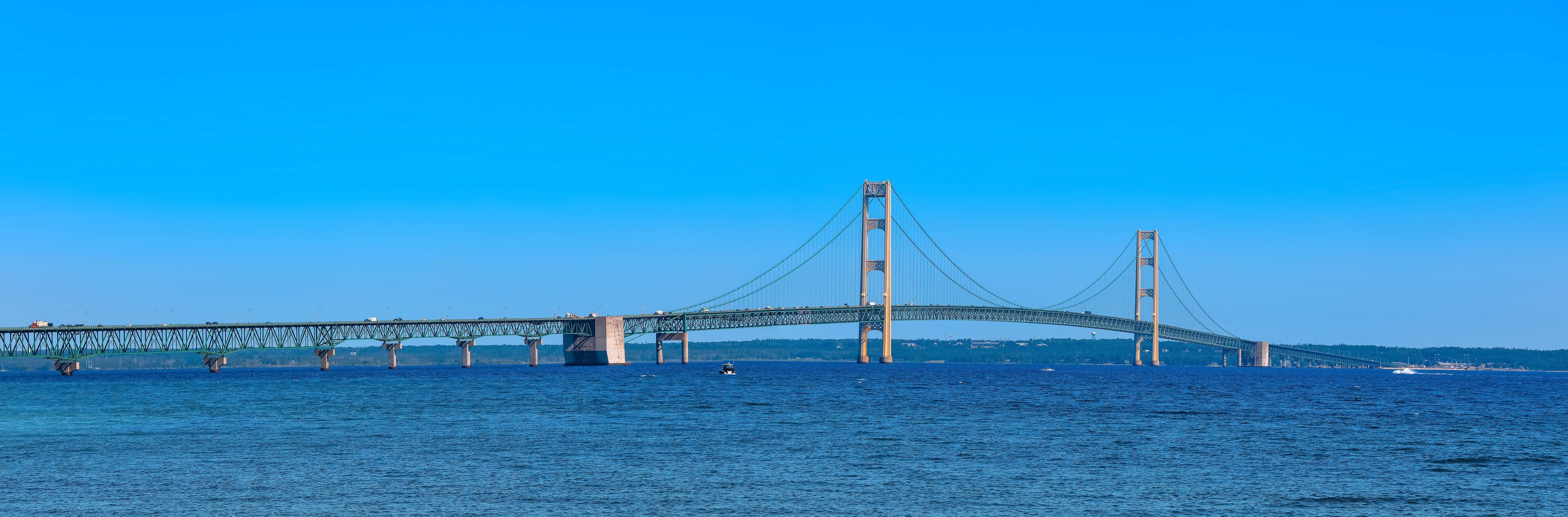 A large suspension bridge spanning a wide river with multiple lanes of traffic and a boat in the water.