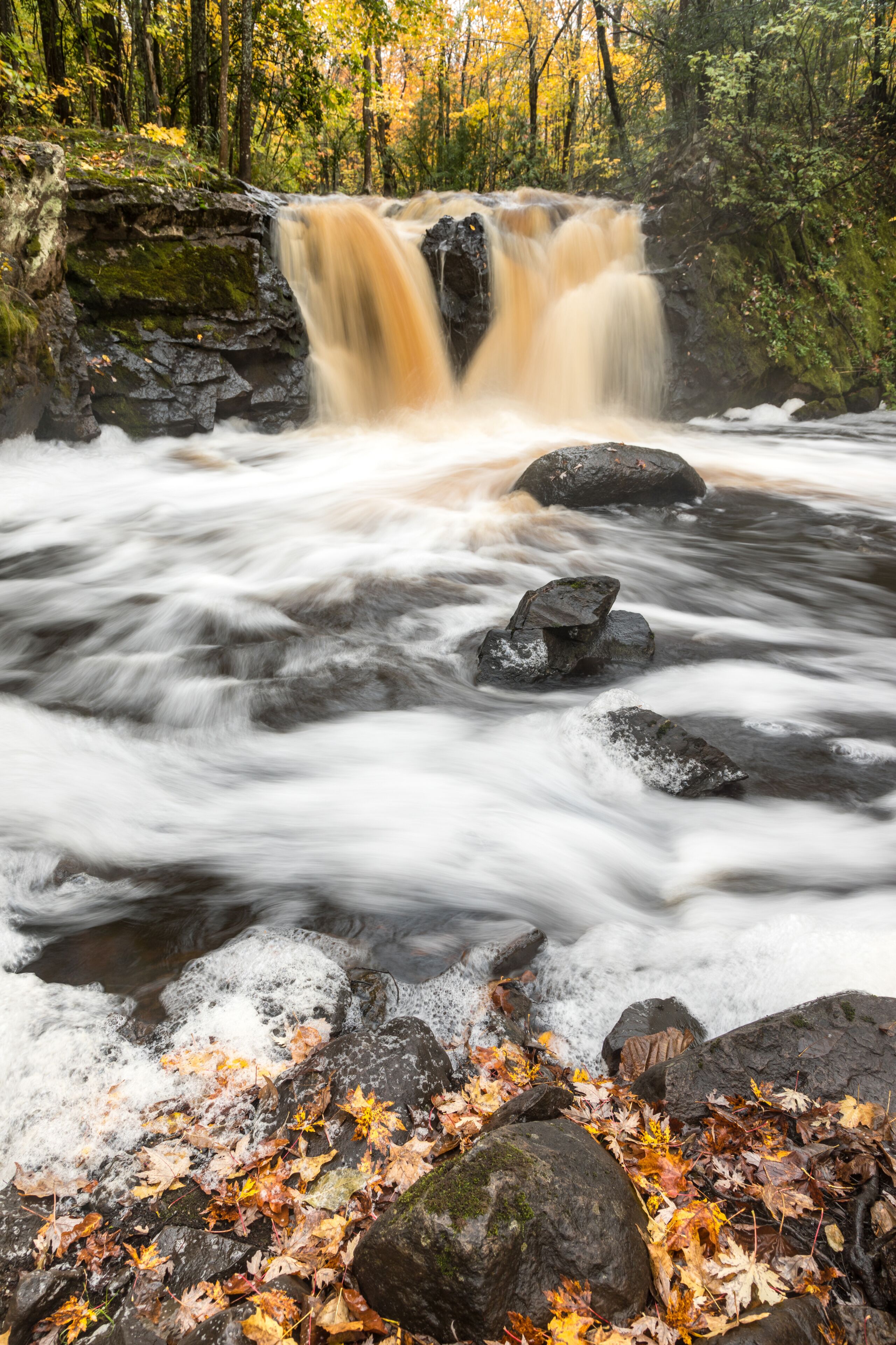 A waterfall cascading over rocks in a forested area with autumn leaves.