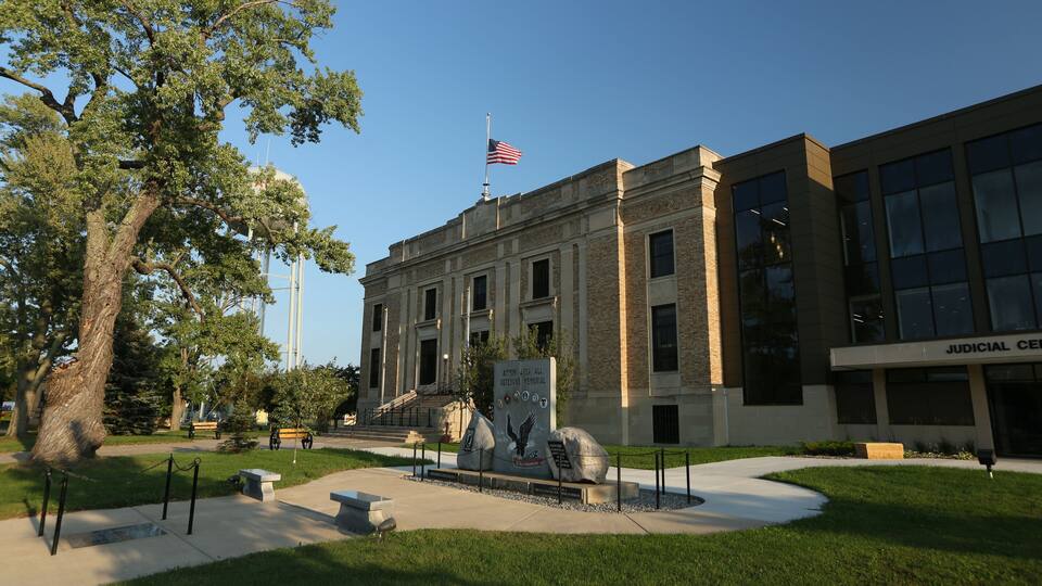 A courthouse with an American flag, a memorial sculpture, and a well-maintained lawn.