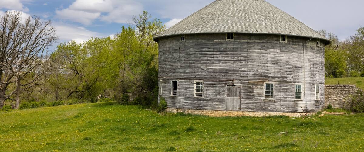A weathered wooden structure with a conical roof, surrounded by a grassy field and trees.
