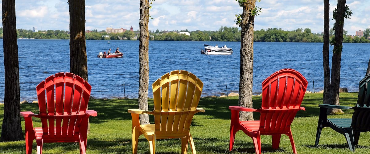 Three Adirondack chairs facing a lake with boats and a distant city skyline.