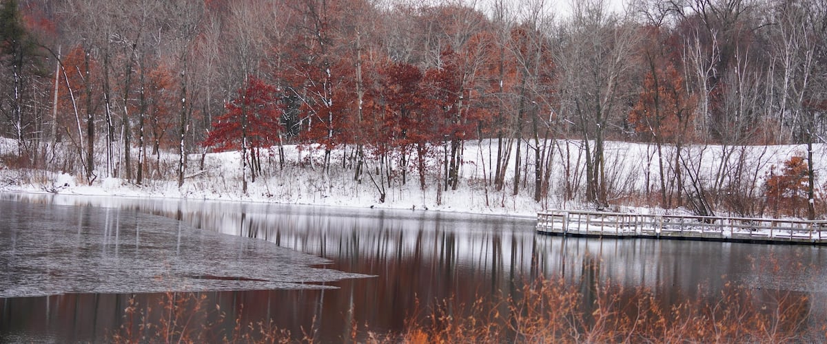 A frozen lake with red autumn leaves, a wooden dock, and a forest in the background.