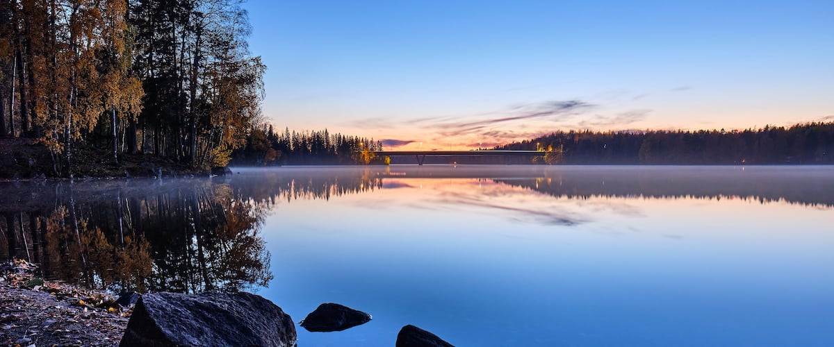 A calm lake at dusk with reflections of trees and a bridge.
