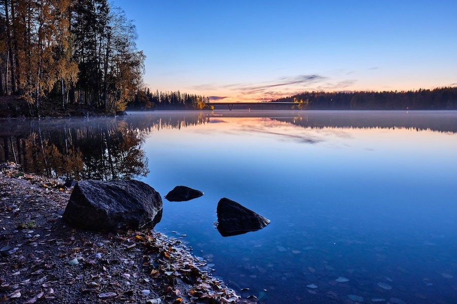 A calm lake at dusk with reflections of trees and a bridge.