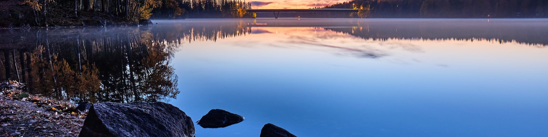 A calm lake at dusk with reflections of trees and a bridge.
