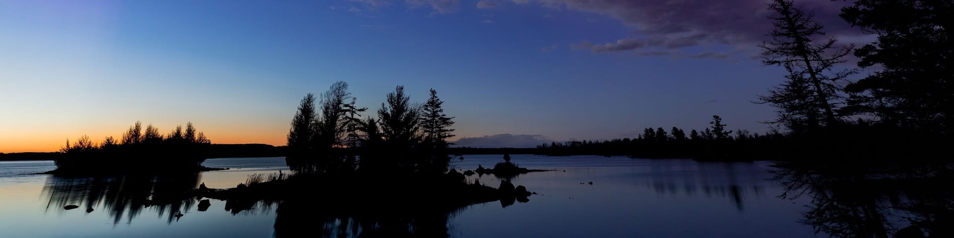 A serene lake at dusk with a reflection of trees, a clear sky, and the Northern Lights.