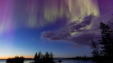 Northern lights erupt over a lake in Minnesota in a dark sky overhead shining rainbow of Aurora light as the sun sets on the horizon