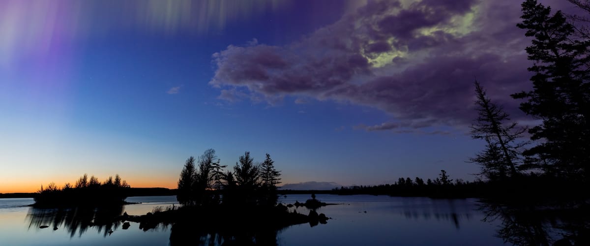 A serene lake at dusk with a reflection of trees, a clear sky, and the Northern Lights.