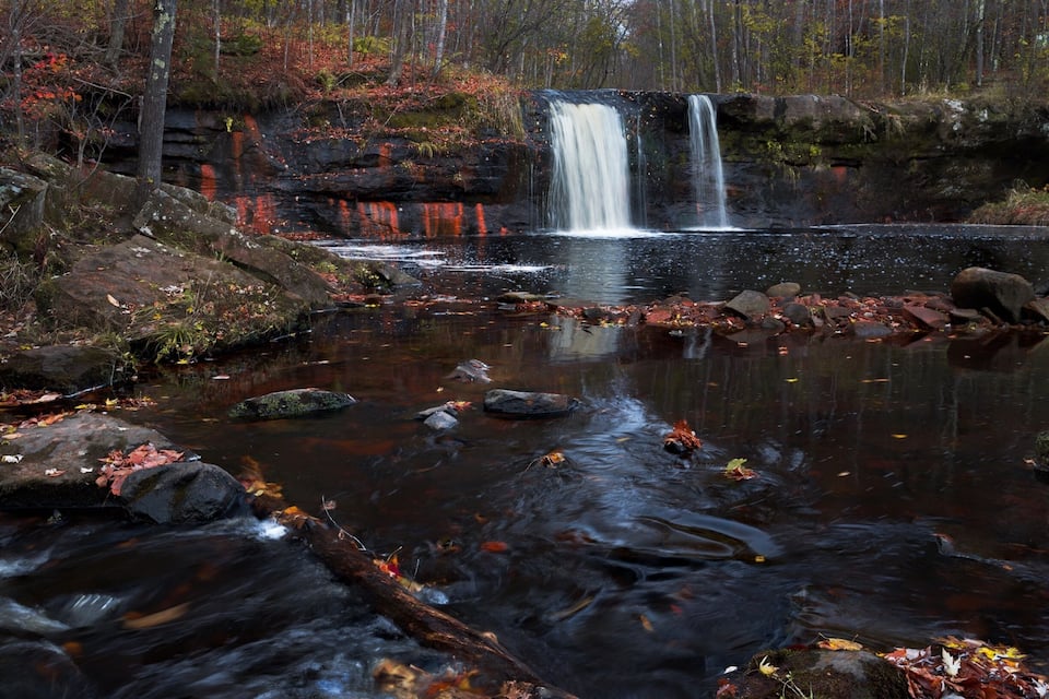 A waterfall cascading over rocks into a forested pond surrounded by autumn foliage.