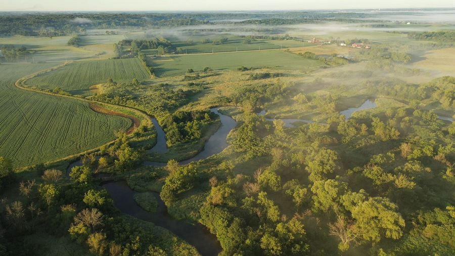 Aerial view of rural landscape with lush greenery, trees, river in the foreground and farmhouse, farmland in the background. American midwestern countryside scenery in the early sunny morning, summer