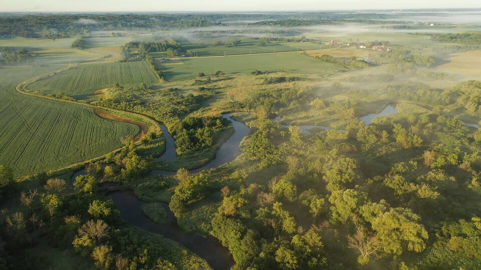 Aerial view of rural landscape with lush greenery, trees, river in the foreground and farmhouse, farmland in the background. American midwestern countryside scenery in the early sunny morning, summer