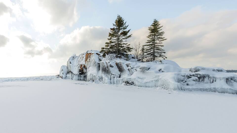 A snowy landscape with icicles hanging from a rock formation and two evergreen trees.