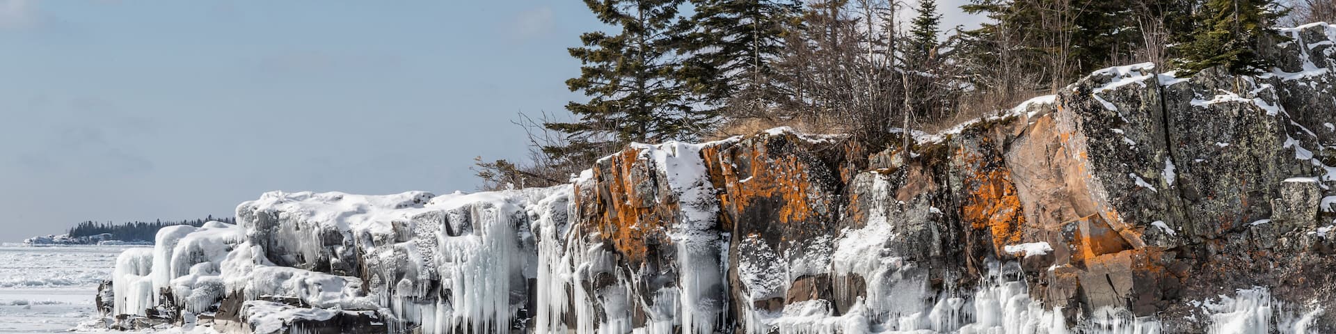 A frozen landscape with icicles hanging from a rocky cliff and snow-covered trees.