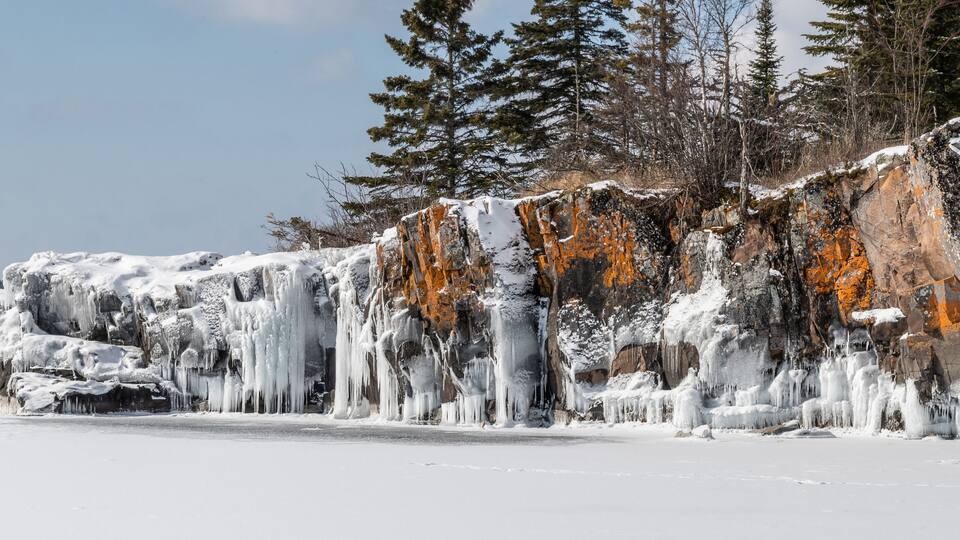 A frozen landscape with icicles hanging from a rocky cliff and snow-covered trees.