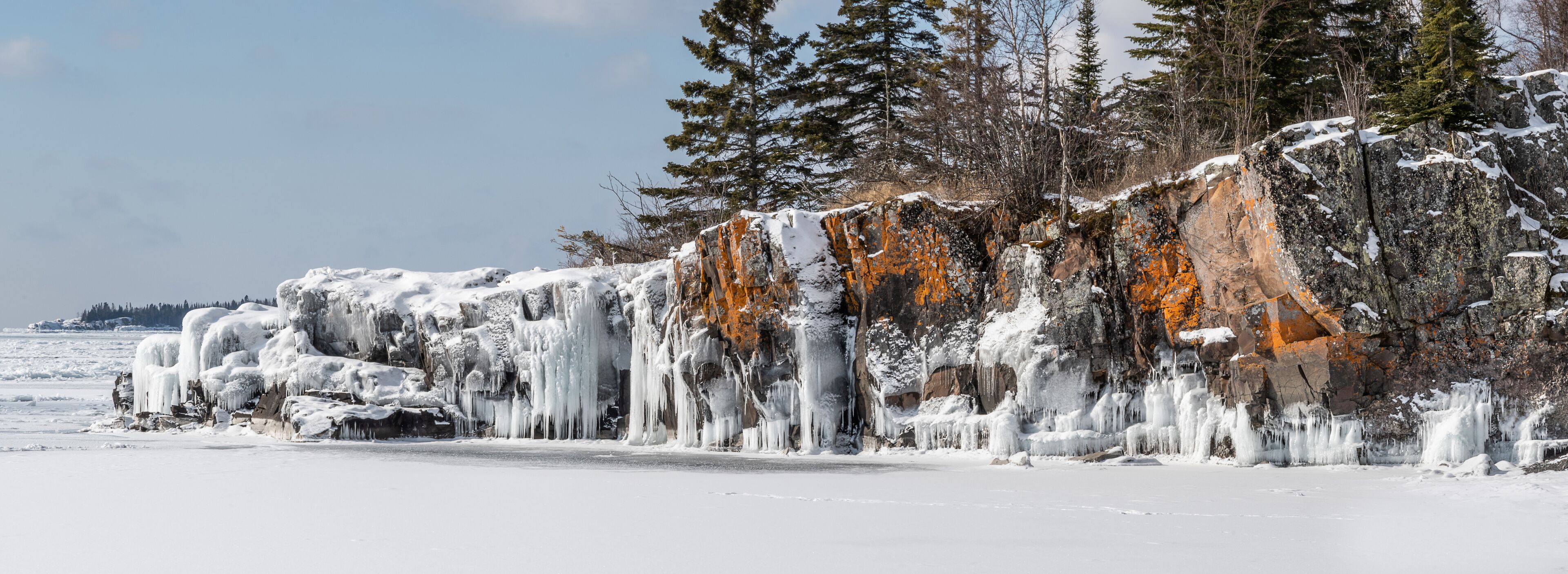 A frozen landscape with icicles hanging from a rocky cliff and snow-covered trees.