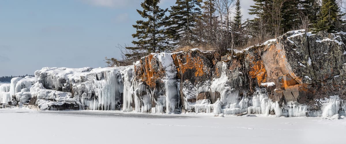 A frozen landscape with icicles hanging from a rocky cliff and snow-covered trees.