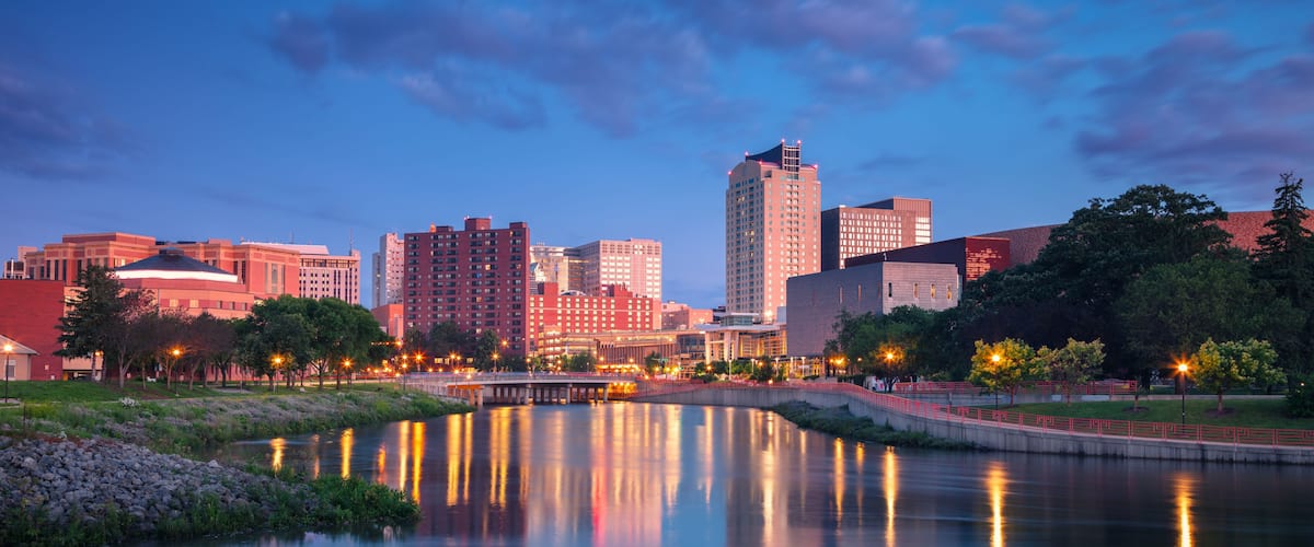 A city skyline at dusk with a river reflecting lights, surrounded by greenery and modern buildings.