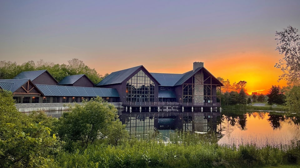 A lakeside lodge with a reflective water surface, surrounded by lush greenery and a sunset sky.