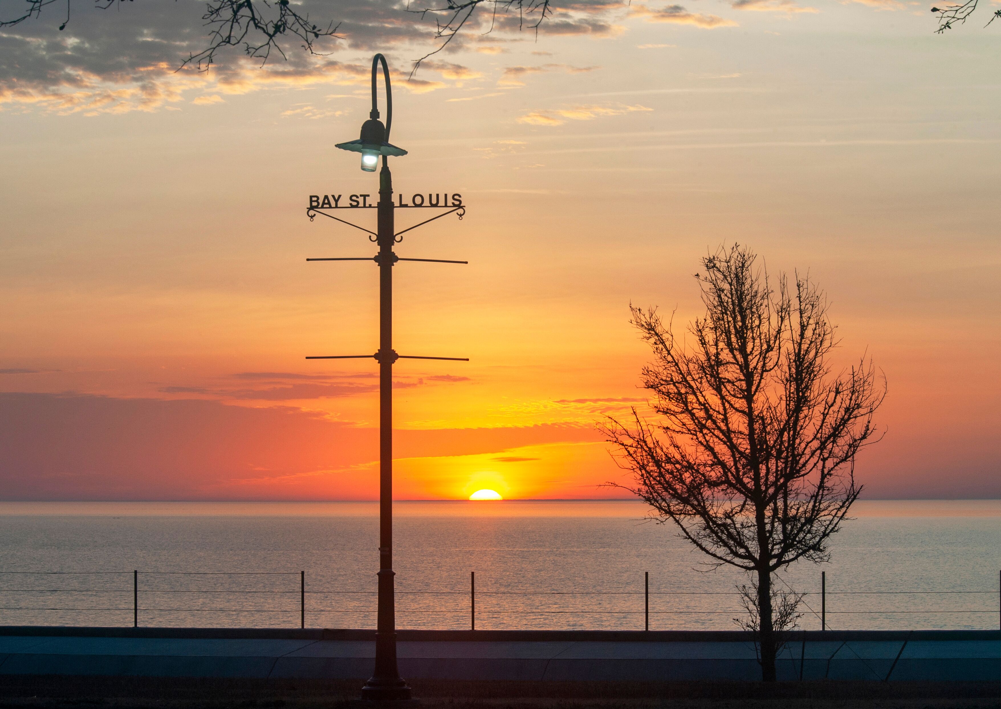 A sunset over the water with a street lamp labeled 'BAY ST. LOUIS' and a leafless tree.