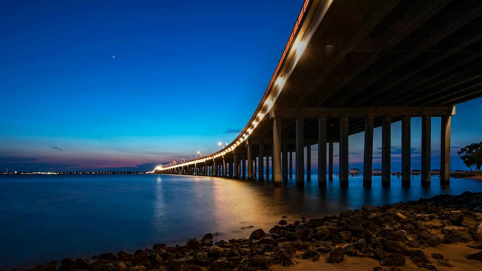 A long bridge extending over calm water at dusk, with a rocky shoreline and a tree-lined path on the right.