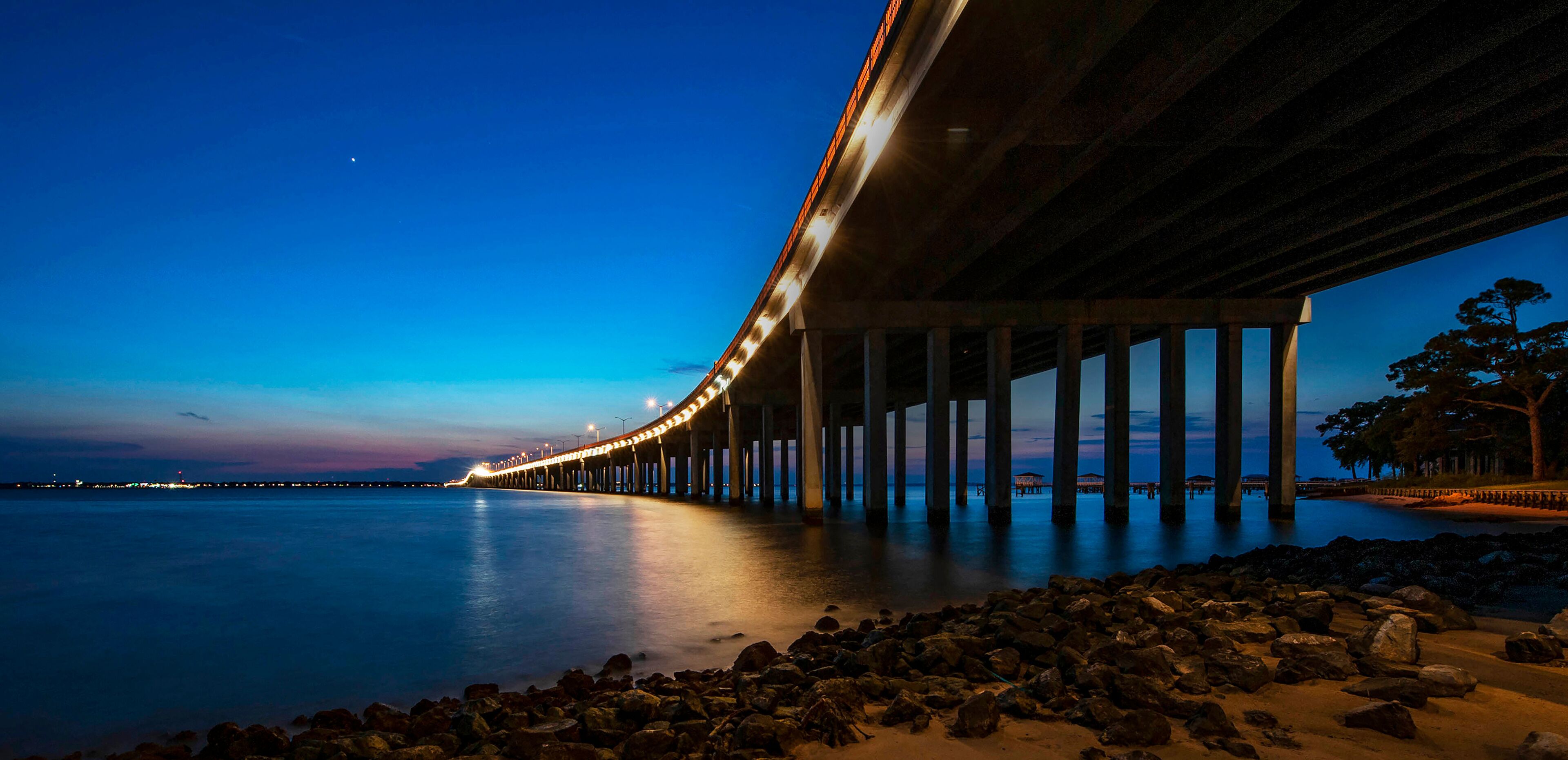 A long bridge extending over calm water at dusk, with a rocky shoreline and a tree-lined path on the right.