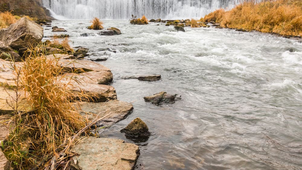 A river with a waterfall in the background, surrounded by rocks and dry grass.