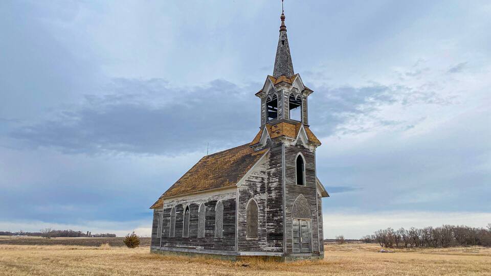 An old wooden church with a tall steeple and arched windows in a grassy field.
