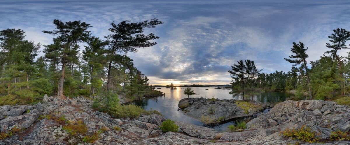 A rocky shoreline with dense coniferous trees and a calm lake reflecting the sky.