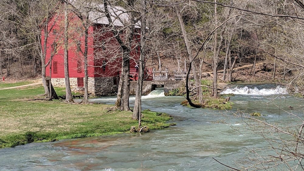Ein rotes Bauernhaus an einem Fluss mit Bäumen im Vordergrund.