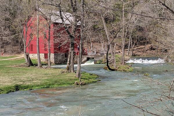 Une grange rouge se dresse près d’une rivière où l’eau coule, avec des arbres au premier plan.