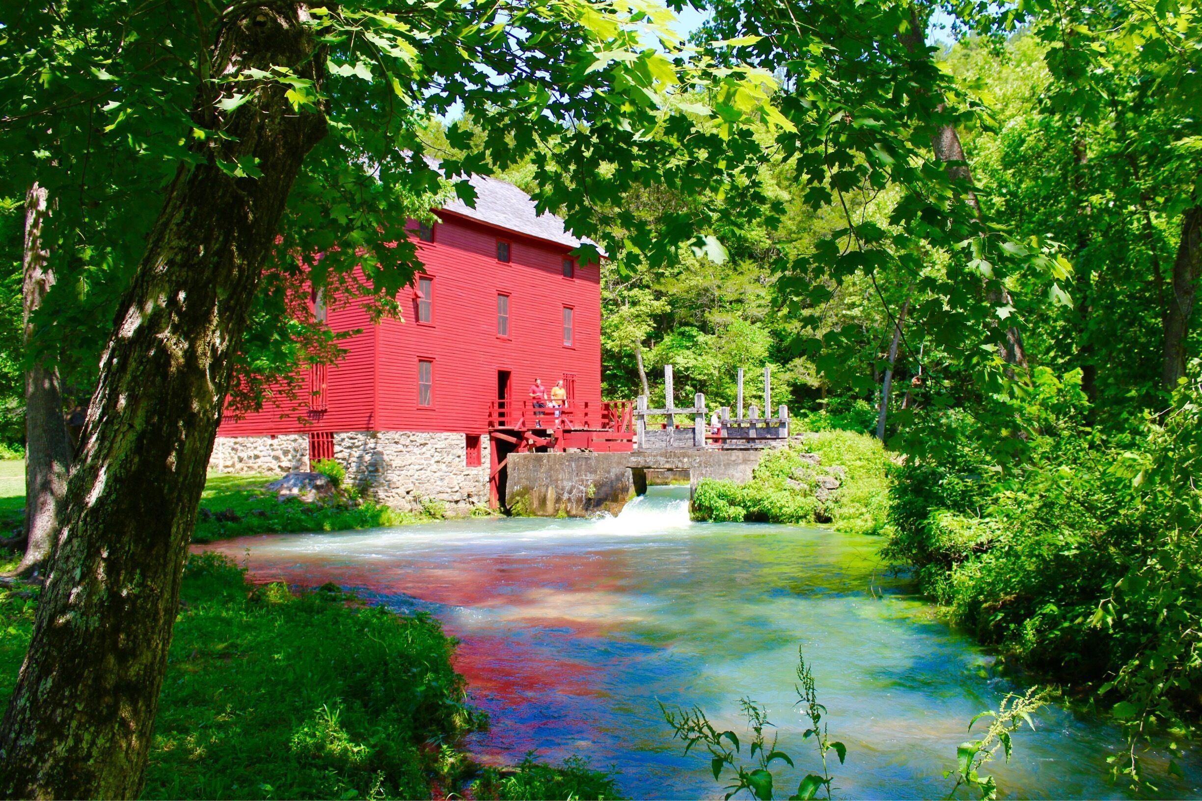 A red mill house by a flowing river, surrounded by lush greenery and a stone bridge.