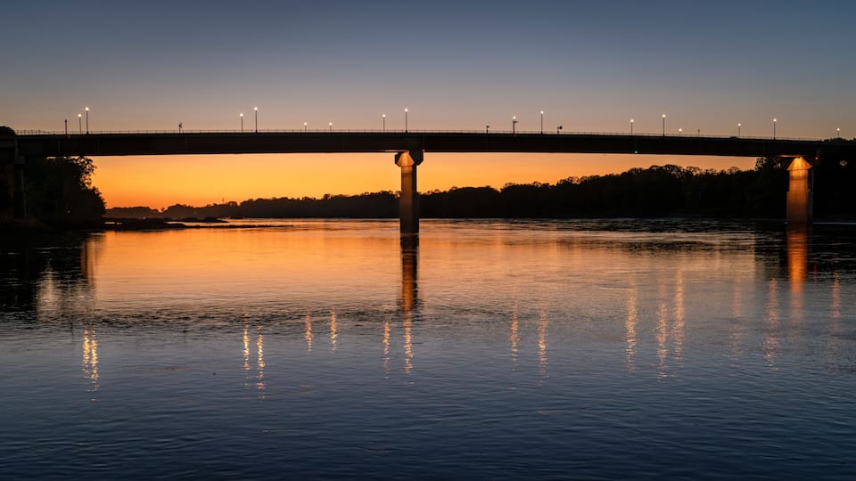 A bridge over a calm river at sunset with lights along the bridge.