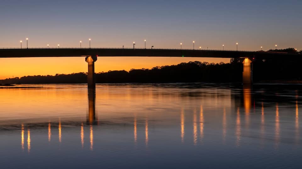 A bridge over a calm river at dusk with streetlights reflecting on the water.