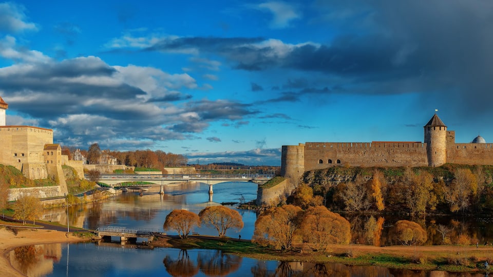 A historic castle with a river in the foreground, a bridge, and autumn foliage.