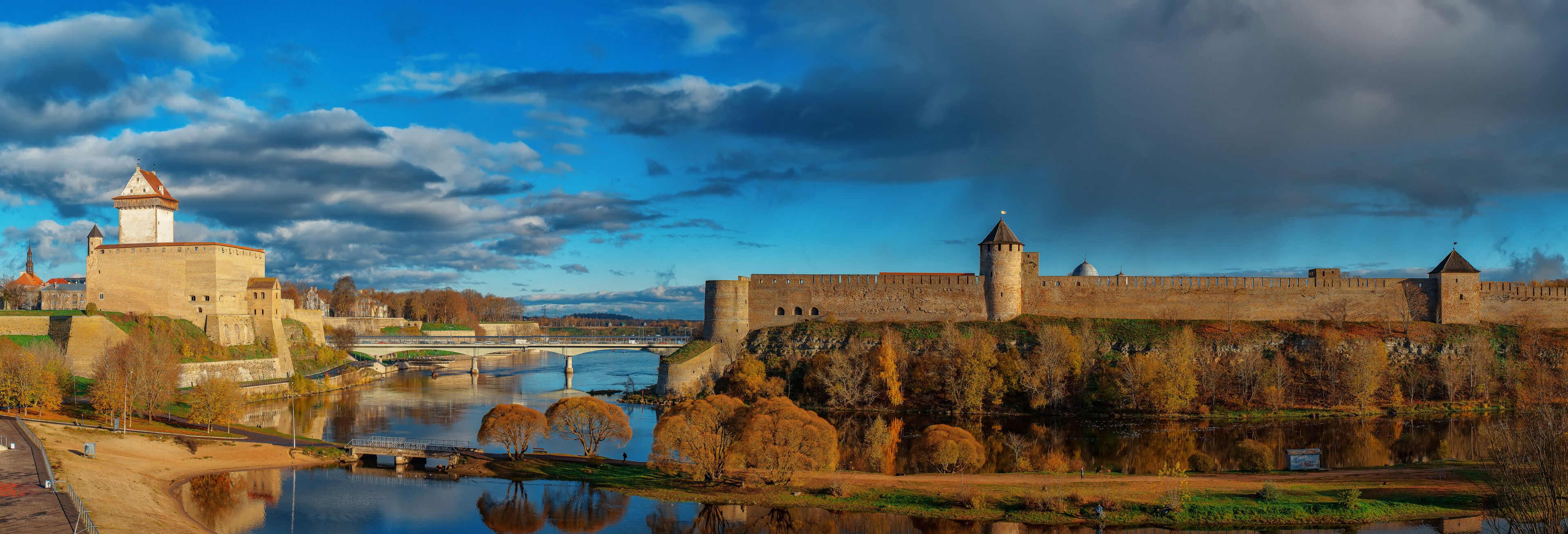 A historic castle with a river in the foreground, a bridge, and autumn foliage.