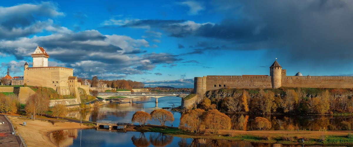 A historic castle with a river in the foreground, a bridge, and autumn foliage.